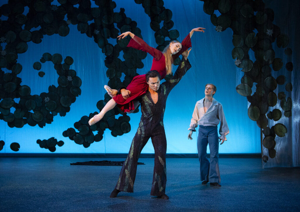 In front of a blue backdrop with dark textured swirls, a male dancer in a dark, scale-y suit lifts a female dancer over his shoulders. She wears a flowing red dress and poses with her back leg in a low retiré derriere, arms lifted. Another dancer stands off to the right with a frightened look on his face. He wears a light colored tunic and blue pants.