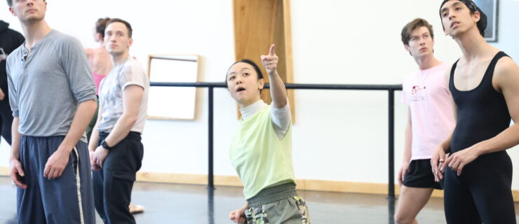 Ching Ching Wong (center) leans back in a slight side lunge and points up to the corner, speaking. Artists of Louisville Ballet surround her and mark her movements.