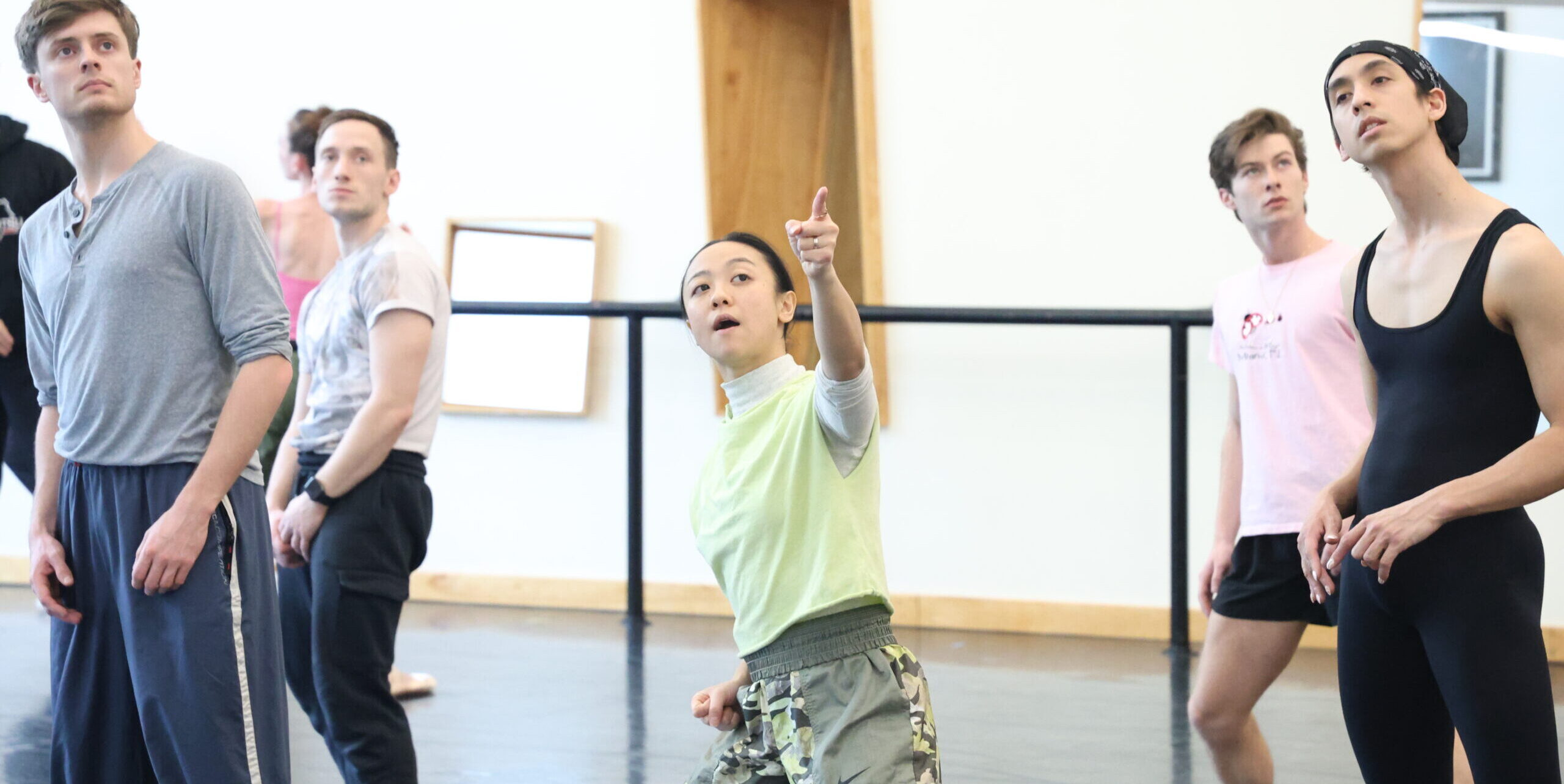 Ching Ching Wong (center) leans back in a slight side lunge and points up to the corner, speaking. Artists of Louisville Ballet surround her and mark her movements.