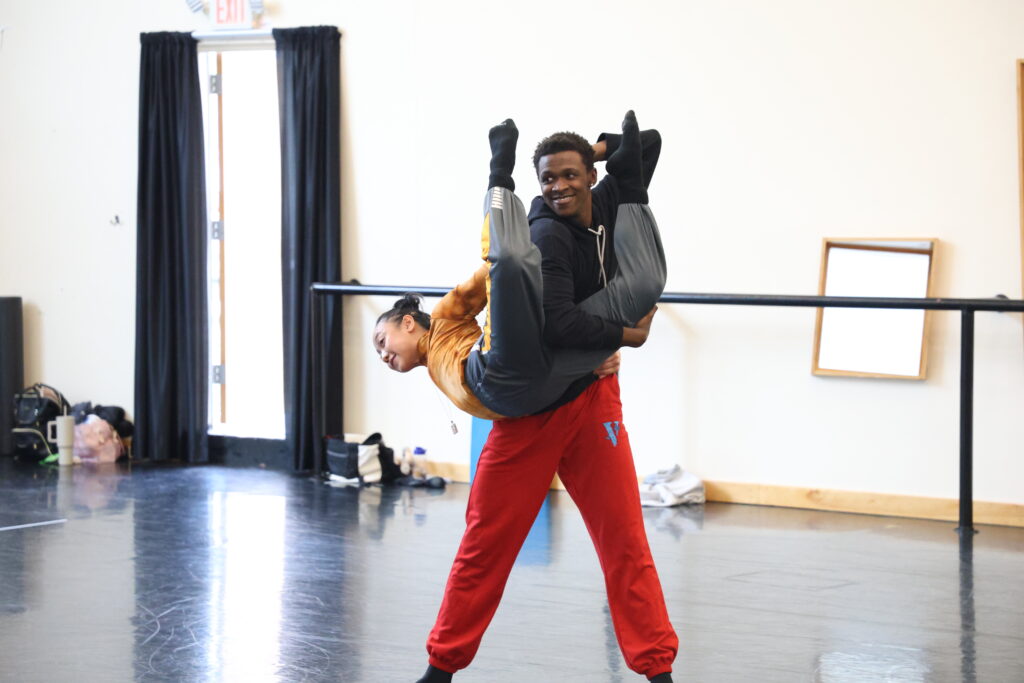 Khalil Jackson, in a black hoodie and red sweatpants, smiles and looks in the mirror as he stands with his torso twisted, holding Ching Ching Wong's right leg in the crook of his right arm. Wong arches back with her legs bent, reaching back with one arm to grab Jackson's other hand behind his back.