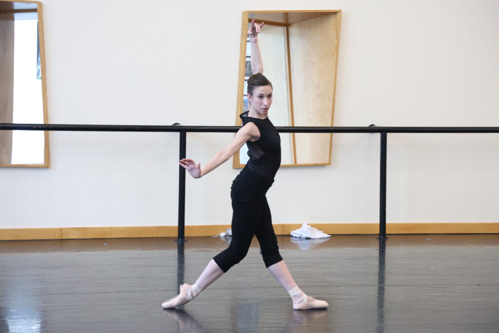 Brienne Wiltsie wears a black leotard and black pants as she rehearses, arms in a stylized high third as she drags her back foot on the floor in a split-leg parallel position.