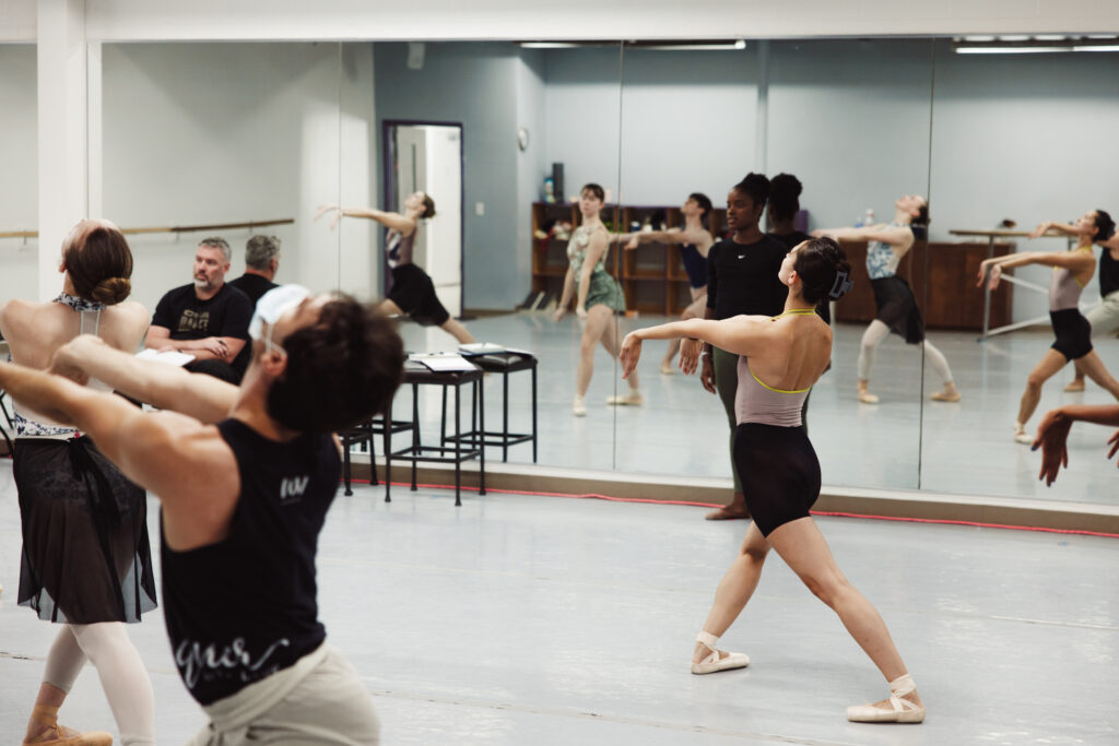 Ingrid SIlva stands with her back to the mirror in a small dance studio and watches a group of dancer as they rehearse. The dancers pose in a wide fourth position in croisé with their arms extended straight out in front of them and their wrists flexed down and their cehsts and eyes lifted up. Silva wears a black shirt and green leggings.