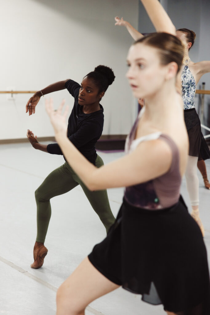 Ingrid SIlva, wearing a black long-sleeved shirt and green leggings, lunges to the side and pops her right foot up onto demi-pointe. She carries her arms to the right side of her torso and makes a circular shape with them. A group of dancers in the foreground and background copy her.