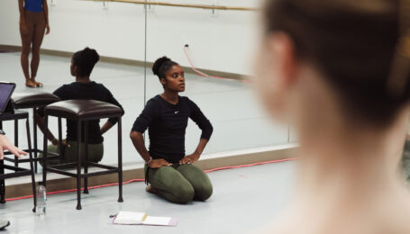 Ingrid SIlva, wearing a long-sleeved black shirt and green leggings, sits on the floor with her knees tucked directly underneath her and her hands on her hips. Her back is towards the mirrors in a dance studio, and she watches a group of dancers in front of her who are mostly out of view of the camera.