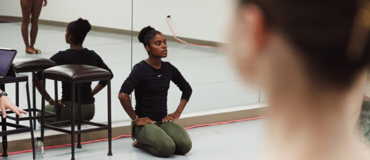 Ingrid SIlva, wearing a long-sleeved black shirt and green leggings, sits on the floor with her knees tucked directly underneath her and her hands on her hips. Her back is towards the mirrors in a dance studio, and she watches a group of dancers in front of her who are mostly out of view of the camera.