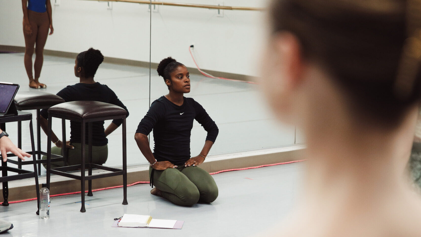 Ingrid SIlva, wearing a long-sleeved black shirt and green leggings, sits on the floor with her knees tucked directly underneath her and her hands on her hips. Her back is towards the mirrors in a dance studio, and she watches a group of dancers in front of her who are mostly out of view of the camera.