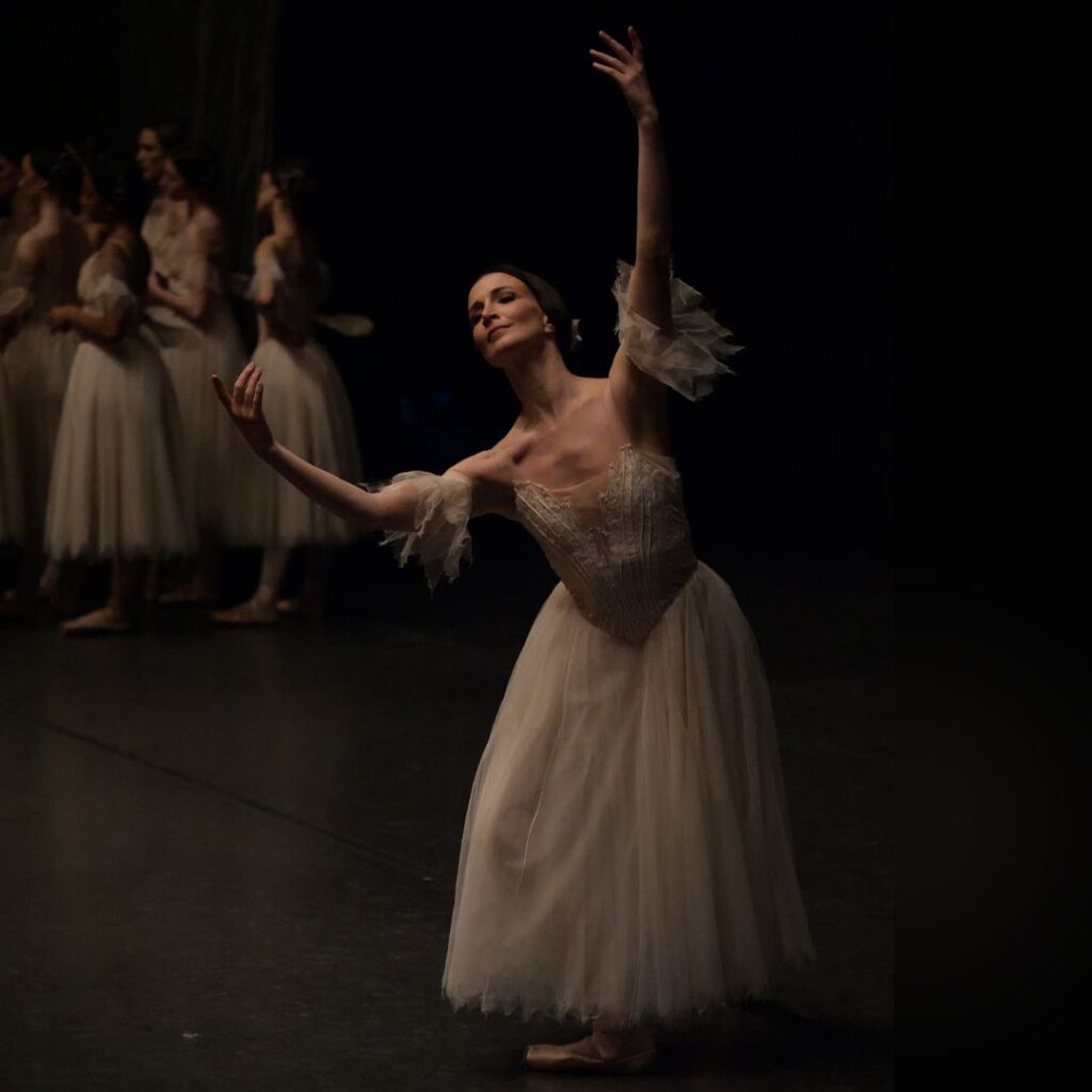 A female dancer dressed in a Romantic-style tutu for "Giselle" practices her port de bras onstage, a peaceful smile on her face, as a group of corps dancers stand together in the back.