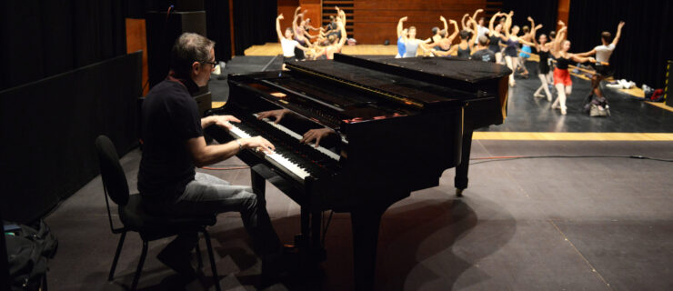 Massimiliano Greco plays on a black piano for a large group of ballet dancers warming up at the barre onstage.