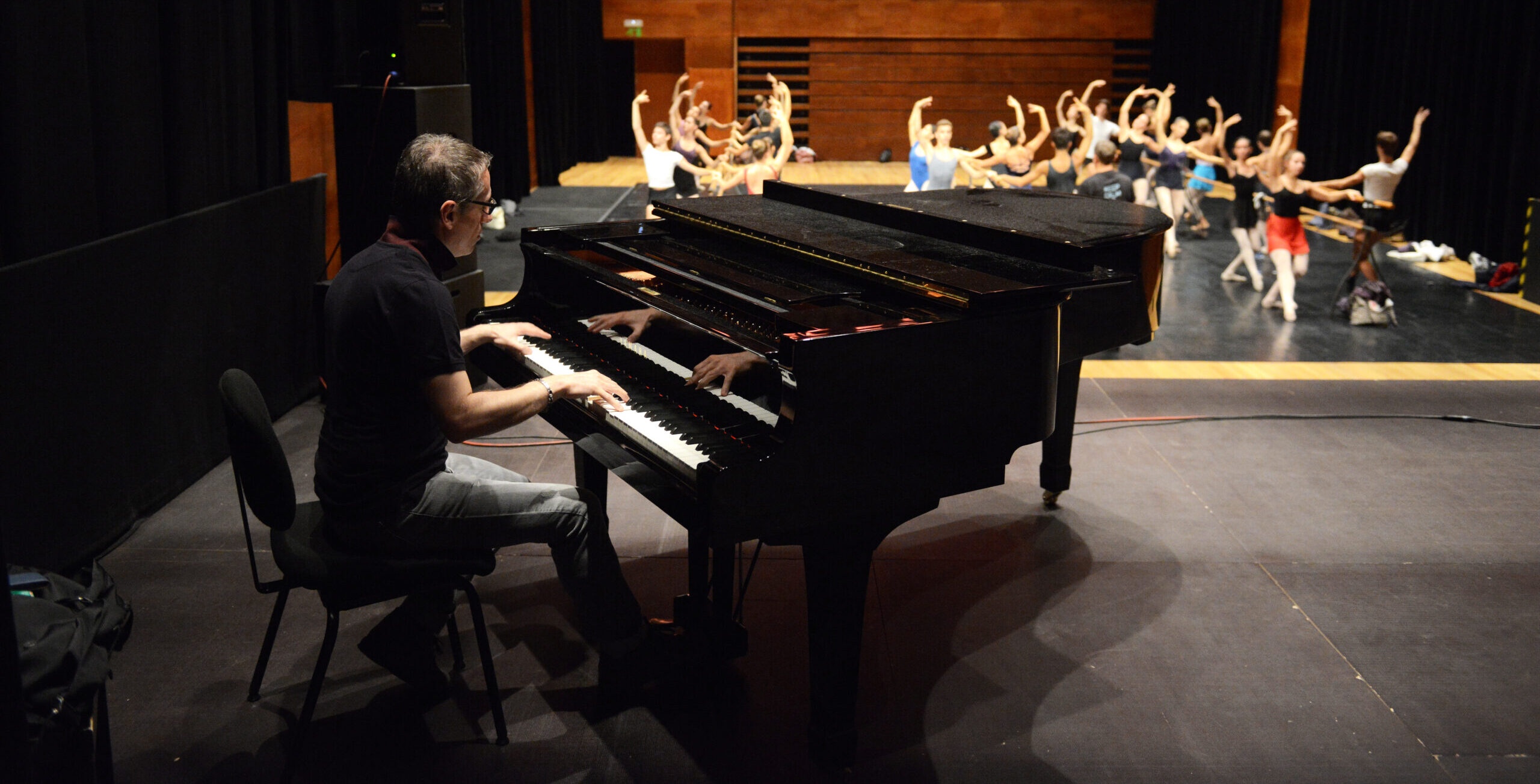 Massimiliano Greco plays on a black piano for a large group of ballet dancers warming up at the barre onstage.