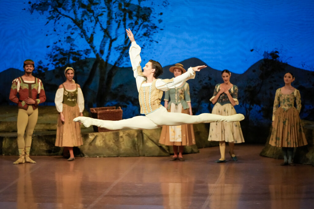 A male dancer does a saut de chat onstage in front of a scenic backdrop onstage as corps members look on in peasant costumes. He wears a puffy-sleeved white shirt under a tan vest, white tights, and white ballet flats.