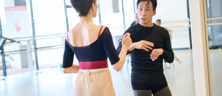 Gen Horiuchi, wear dance practice clothes, talks to a ballerina during a rehearsal in a brightly lit dance studio.