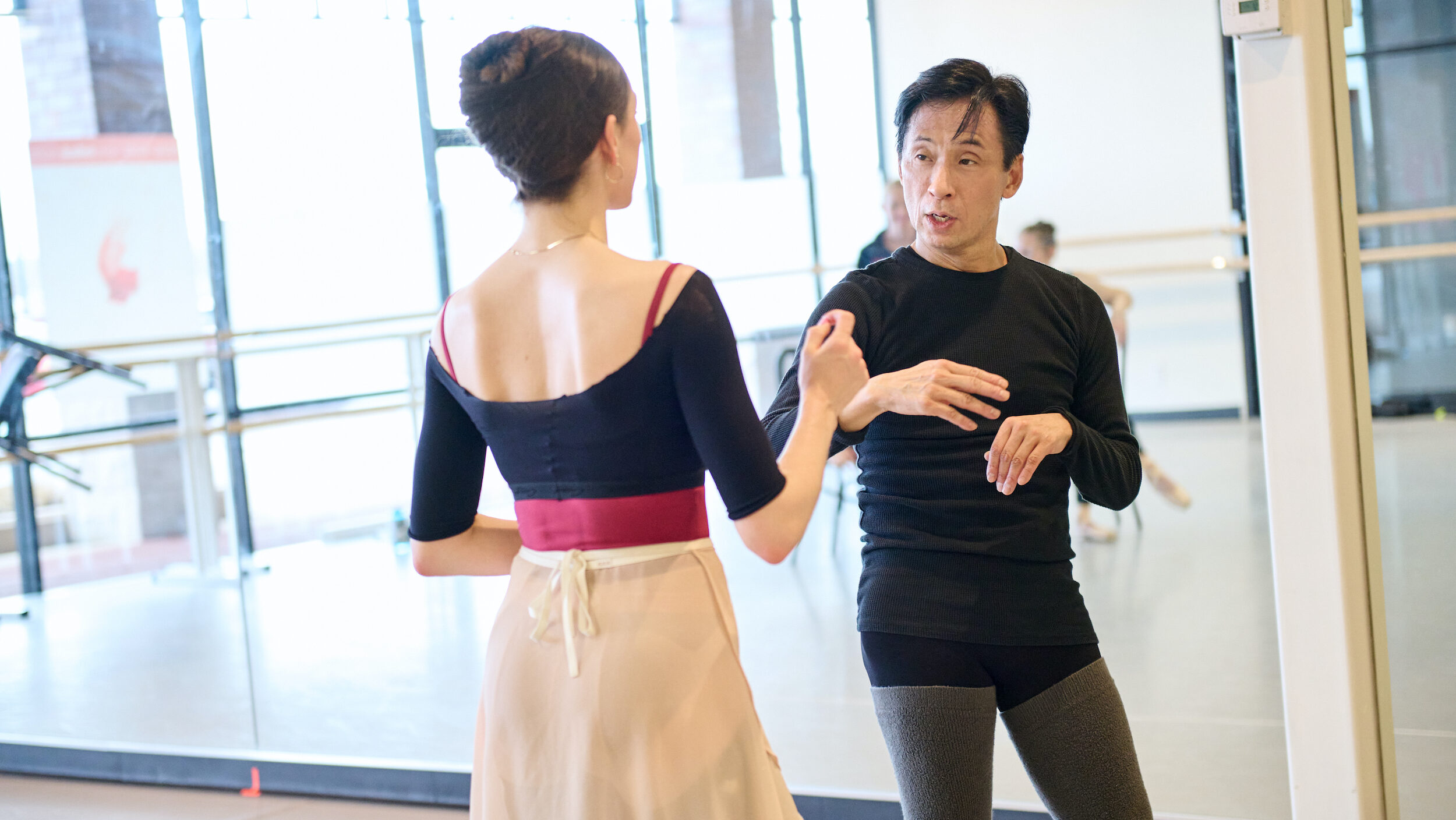 Gen Horiuchi, wear dance practice clothes, talks to a ballerina during a rehearsal in a brightly lit dance studio.