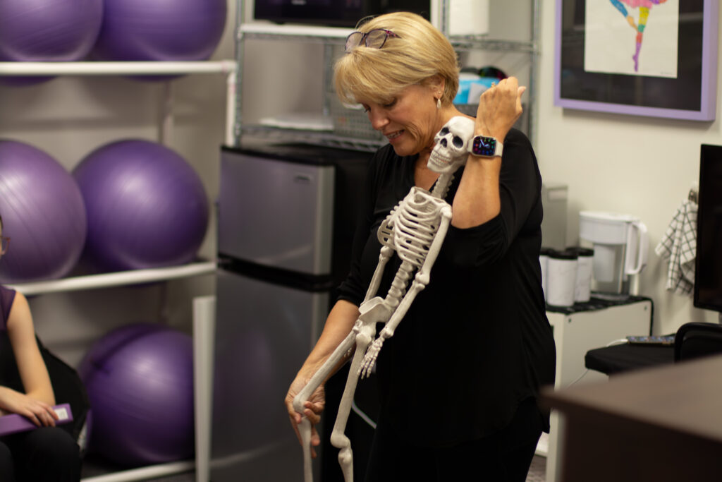 Gretchen Vogelzang, wearing a black shirt and pants, holds a small model skeleton against her body as she talks to her ballet students (off camera). She looks down at the skeleton and bends its right knee with her right hand.