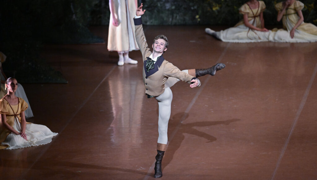 A male ballet dancer poses in attitude derriere on stage, beaming with his arms in high third. He wears a period-specific tan jacket with a shiny gray bow at the neck, white tights, and black dance boots.