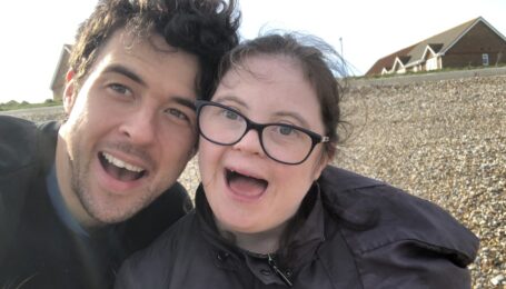 Thomas Forster stands very close to Louise Forster, theier heads touching as they take a selfie with big, open-mouthed smiles. They are outside and wearing jackets, with a road and houses in the background.