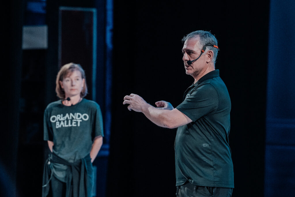 Jordan Morris is shown from the waist up wearing a headset and rehearsing dancers (not shown) onstage. A woman in an Orlando Ballet t-shirt stands off to the side and watches him.