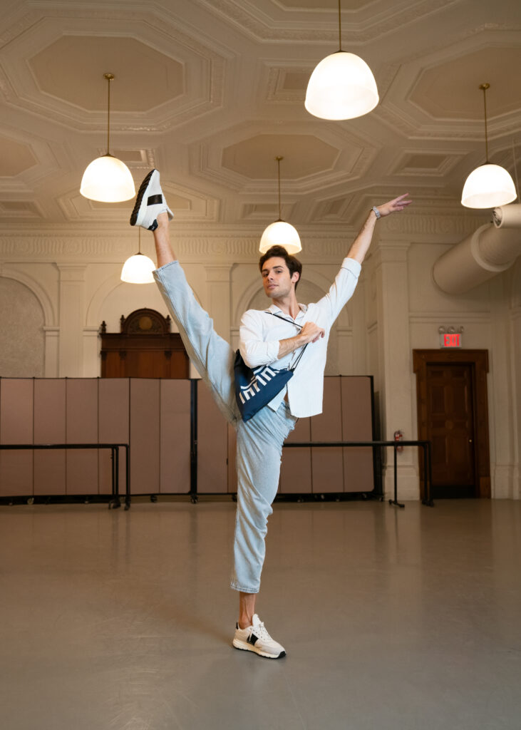 Jacopo Tissi developpes his right leg while holding his dance bag. He wears a watch on his left wrist.