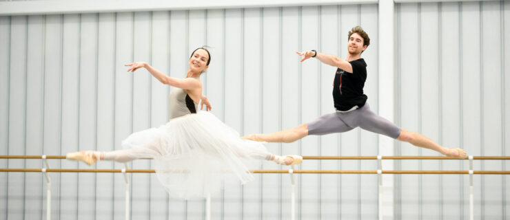 Katja Khaniukova and Aitor Arrieta wear dancewear and leap through the air side-by-side in a large dance studio. Khaniukova also wears a long, white practice tutu.