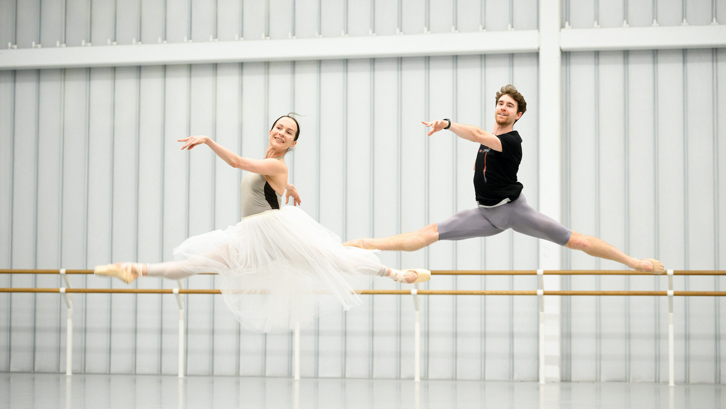 Katja Khaniukova and Aitor Arrieta wear dancewear and leap through the air side-by-side in a large dance studio. Khaniukova also wears a long, white practice tutu.