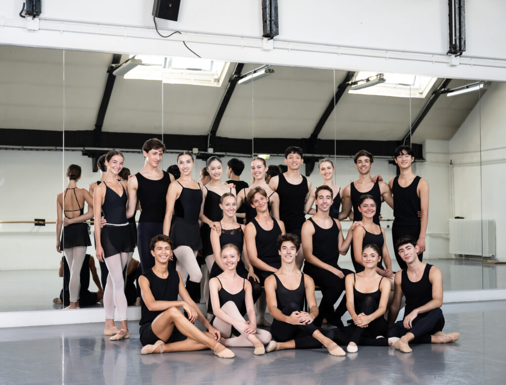 18 young male and female dancers in black ballet class attire pose for a group photo in a large ballet studio, with their backs to the mirror.