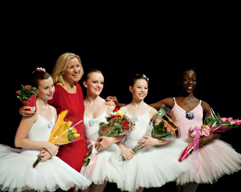 Dayna Fox, wearing a red dress, stands next to a group of young ballet dancers in white or pink tutus. They wrap their arms around each others' shoulders and smile. The young dancers hold bouquets of flowers.