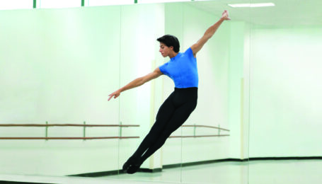 A teenage male ballet student wearing black tights over a bright blue leotard does a brisé volé in a dance studio.