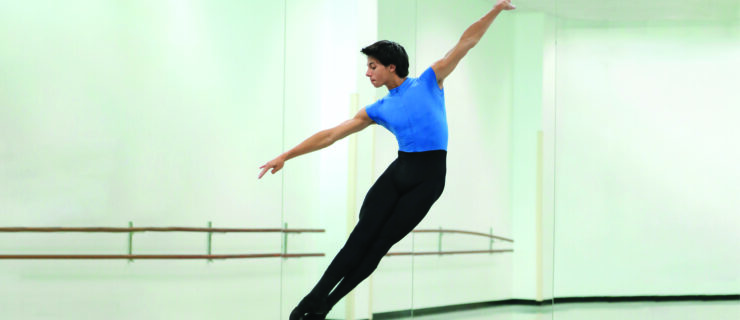 A teenage male ballet student wearing black tights over a bright blue leotard does a brisé volé in a dance studio.