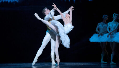 Jurgita Dronina, as Odette in a white feathered tutu, balances on pointe in a high arabesque as she arches backwards. Harrison James, as Siegfried, holds her wrists. Her arms are in a V overhead.