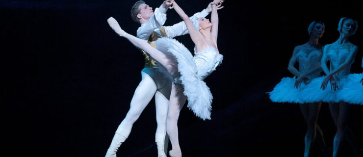 Jurgita Dronina, as Odette in a white feathered tutu, balances on pointe in a high arabesque as she arches backwards. Harrison James, as Siegfried, holds her wrists. Her arms are in a V overhead.