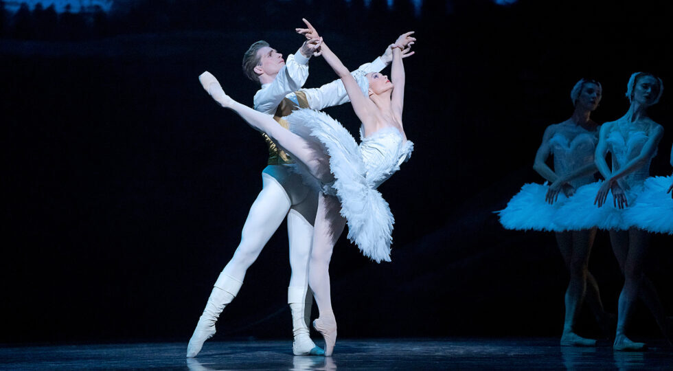 Jurgita Dronina, as Odette in a white feathered tutu, balances on pointe in a high arabesque as she arches backwards. Harrison James, as Siegfried, holds her wrists. Her arms are in a V overhead.