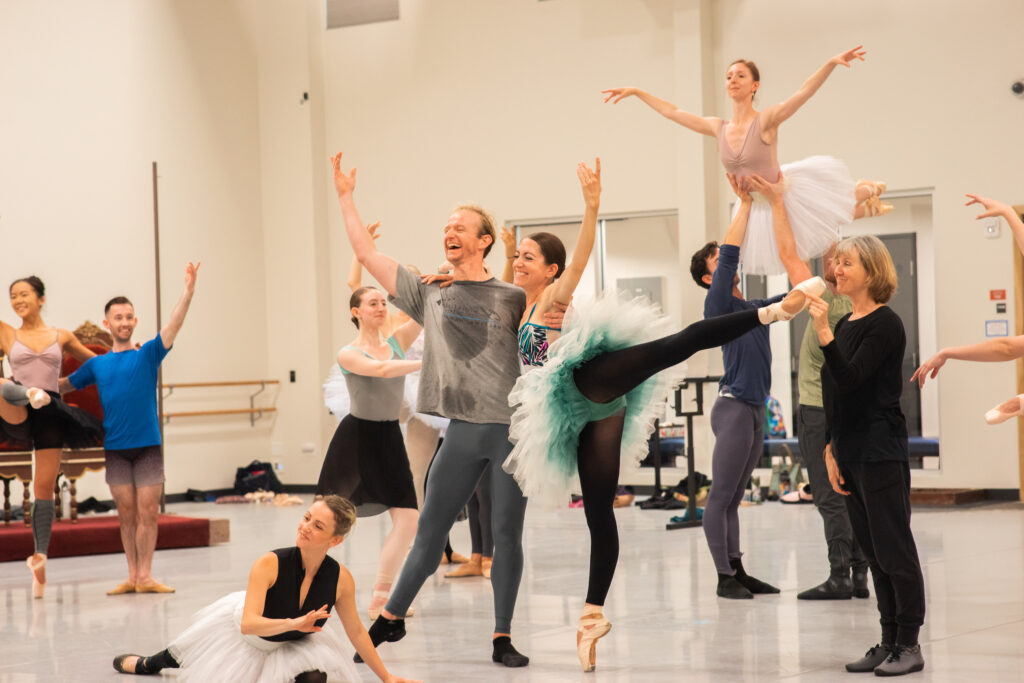 A group of professional ballet dancers pose in a tableau during rehearsals. The women wear practice tutus and one is held aloft by two men in the back as a couple poses up front.