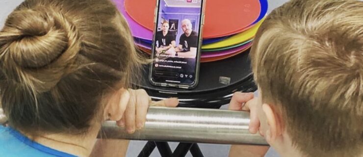 A boy and girl in dance clothing are shown from behind. They lean their faces against a barre and look at a phone propped up in front of them, where two ballet teachers talk to them.