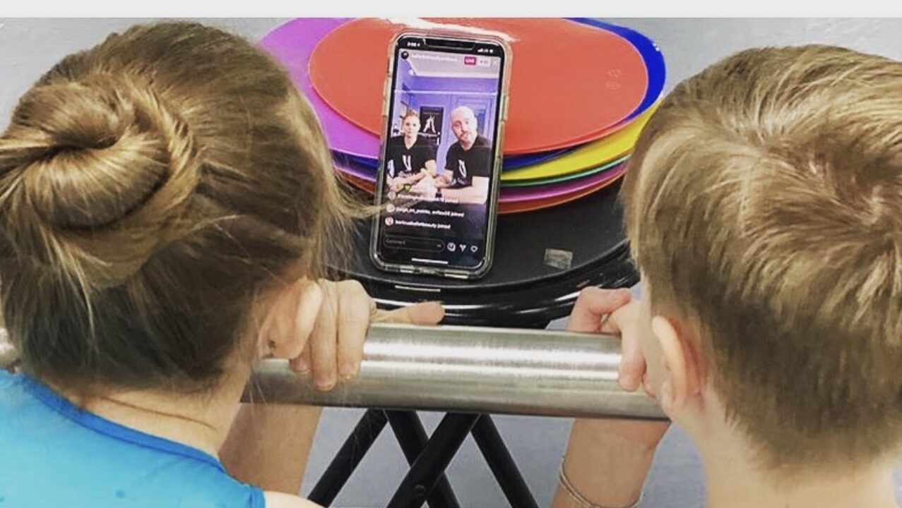 A boy and girl in dance clothing are shown from behind. They lean their faces against a barre and look at a phone propped up in front of them, where two ballet teachers talk to them.