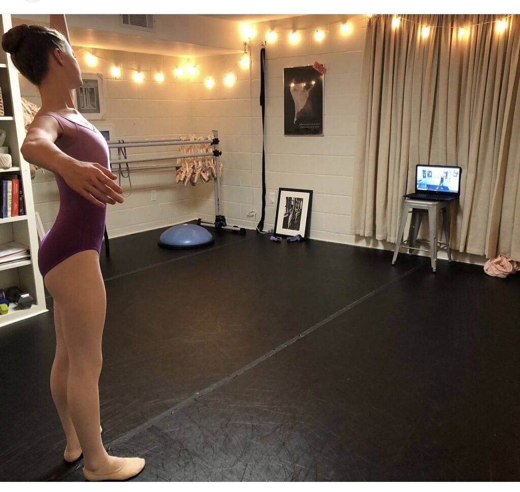 A young teenage girl in a small dance studio practices her port de bras while watching a virtual ballet class on a laptop propped up on stool.