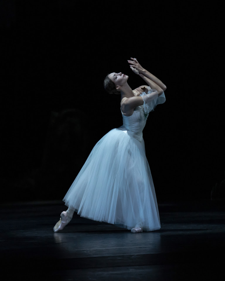 A female dancer in a long white Romantic tutu performs onstage as "Giselle." She stands in tendu derriere facing profile and arches up to look at the sky, her arms raised and bent so her hands appear to be asking or praying.