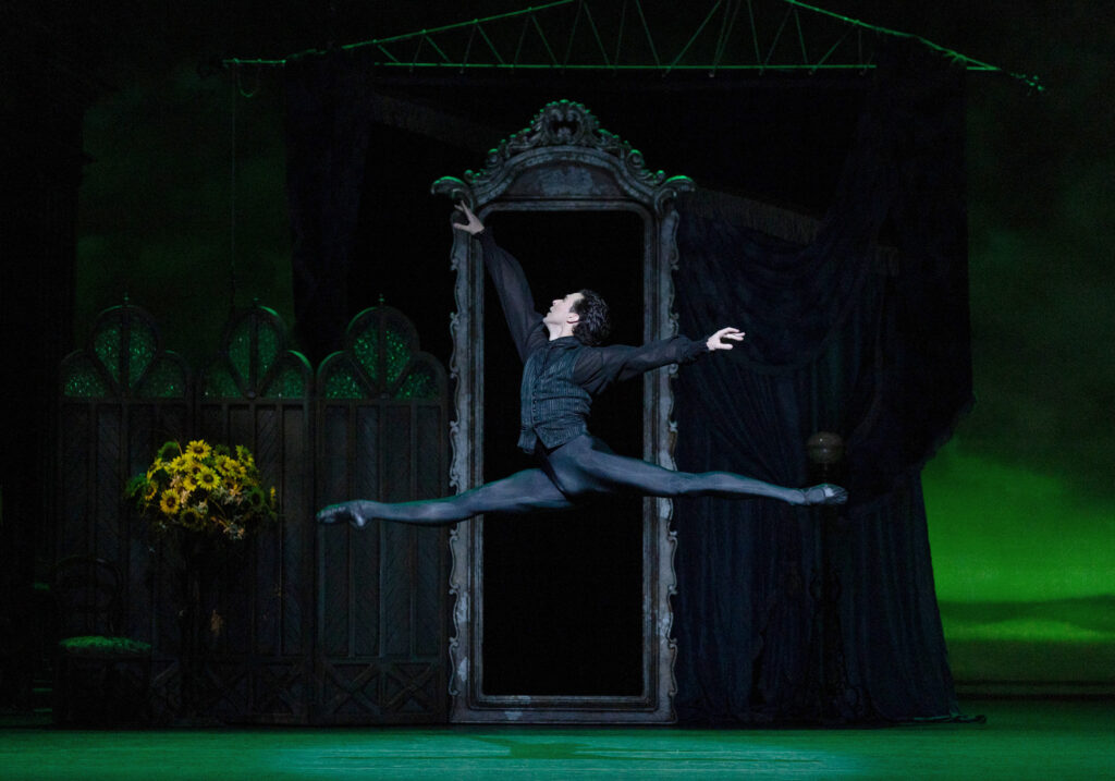 Thomas Forster, dressed in black tights and ballet slippers and a black jacket, does a saut de chat onstage during a performance. His arms are in first arabesque, and he danes in front of a large set with a floor-to-ceiling mirror in the background.