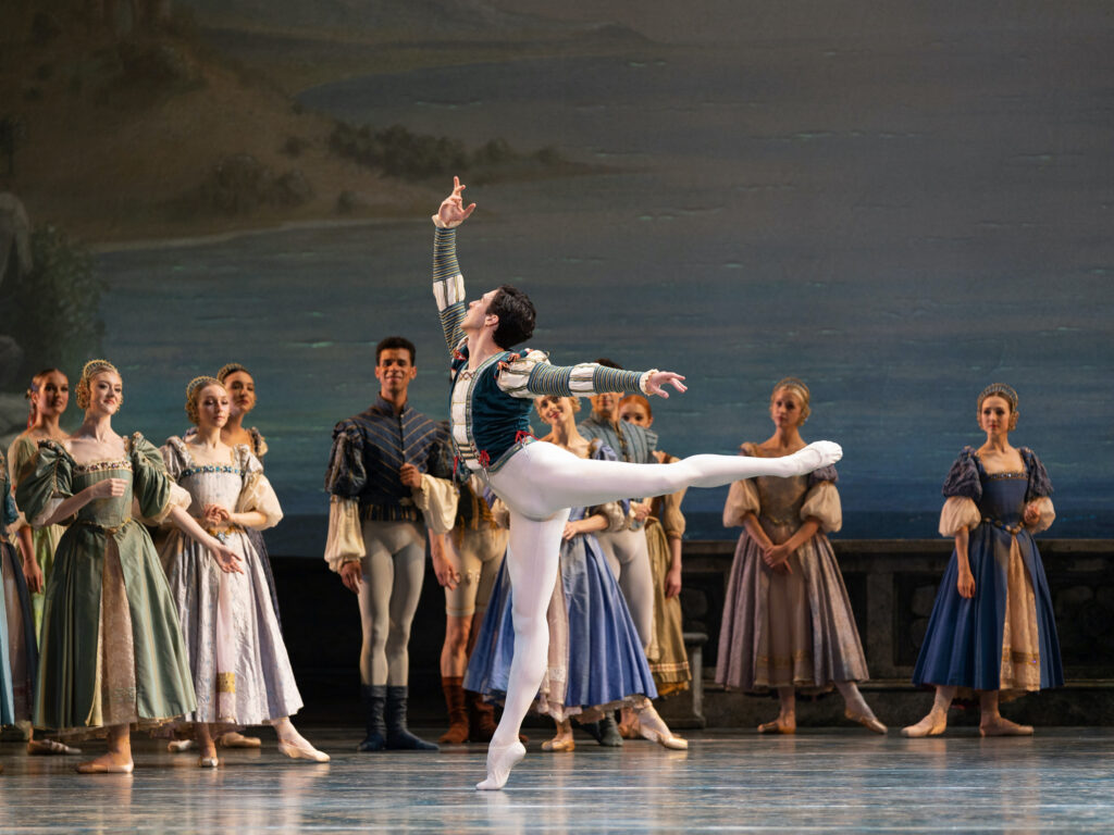 Thomas Forster, wearing white tights and a green and white tunic, does an arabesque facing stage right during a performance of Swan Lake. A corps of dancers costumed as noble men and women stand in the background and watch him.