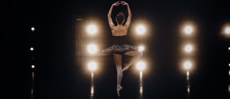 A young ballerina in a black tutu is shown from the back in a passé on pointe, her arms in fifth position en haut. She faces the wings onstage, with rows of the brightly lit booms.