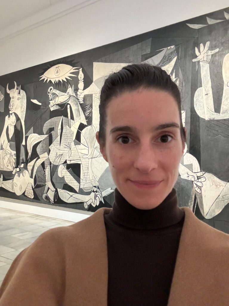 A young woman in a brown turtleneck and camel-colored coat takes a selfie in front of a large, black and white cubist mural while visiting an art museum.