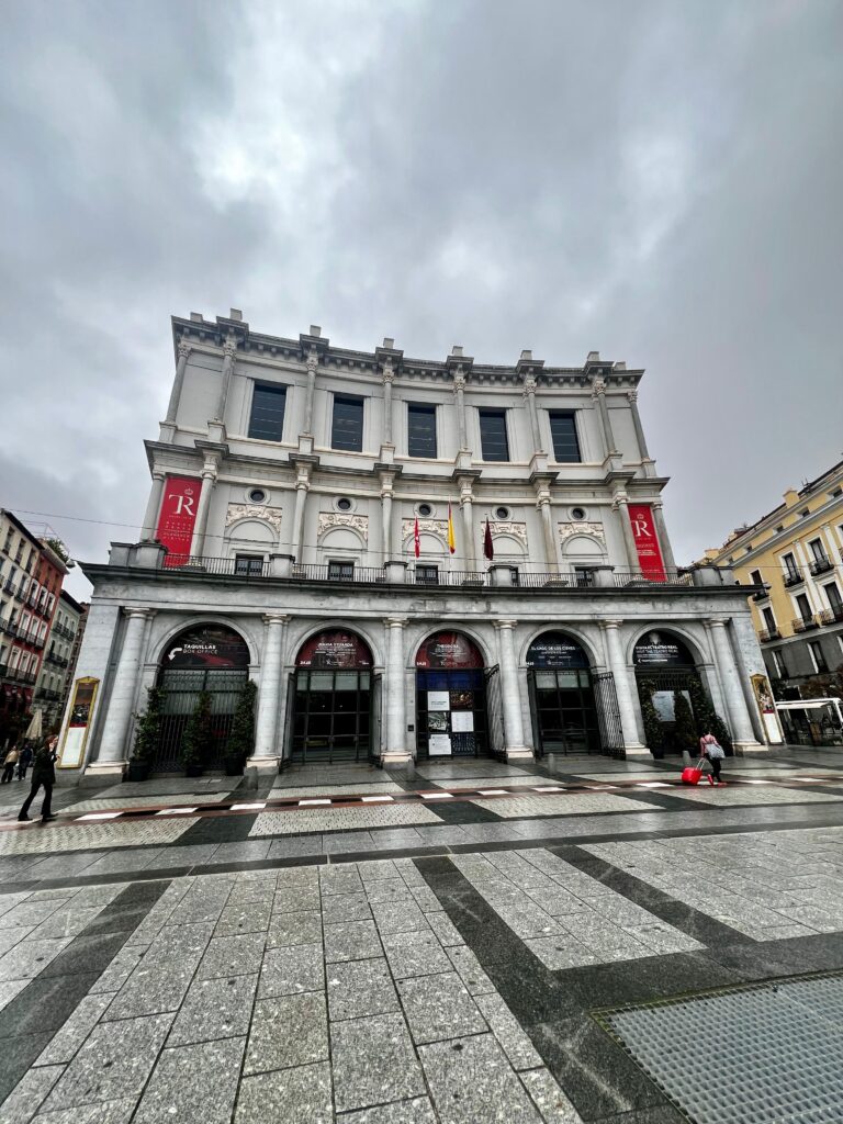 A photo of a theater in a city square made of stone and with pillars and archways at its entrance.