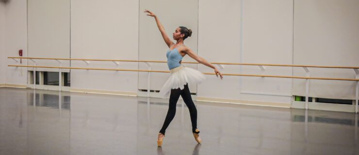 Kimbrough, a Black ballerina, stands en pointe in a dance studio, wearing a blue leotard, black tights, a white tutu and nude pointe shoes.