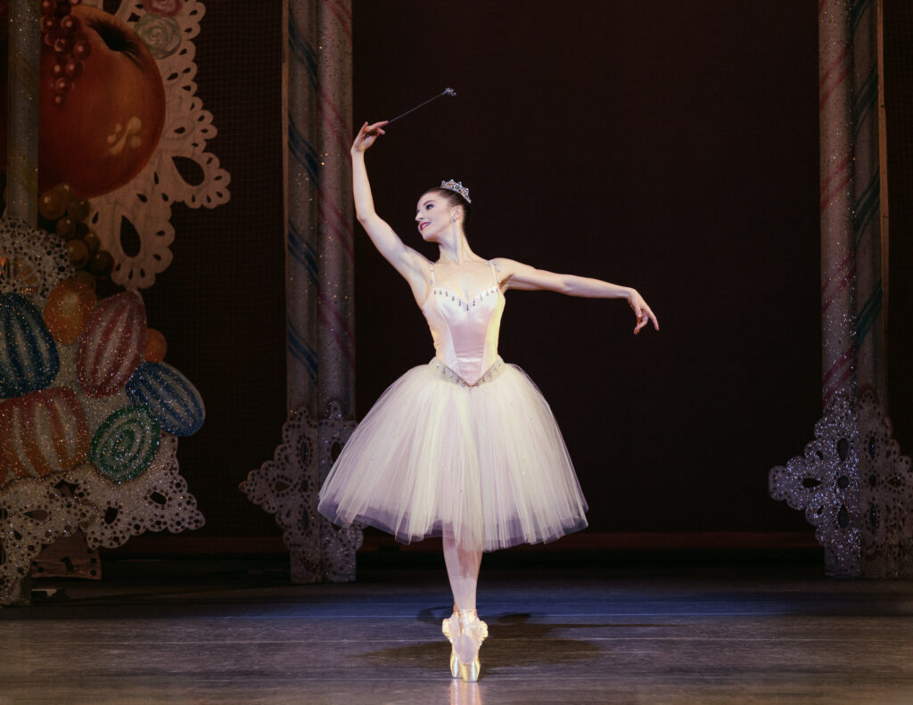 Indianan Woodward stands in sus-sous on pointe with her right arm lifted and her left arm out to the side during a performance. She wears a knee-length pink tutu with a pink satin bodice, and holds a silver wand in her right hand.