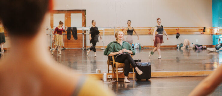 Claire Kretzschmar sits in a chair at the front of a ballet studio and laughs as she leads rehearsal, surrounded by female ballet dancers.