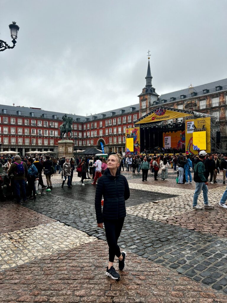 A young woman with blonde hair wears a black long-sleeved shirt, leggings and sneakers and stands in the middle of a busy cobblestone square in Spain. She crosses her right foot over her left and looks up towards her right, smiling slightly.