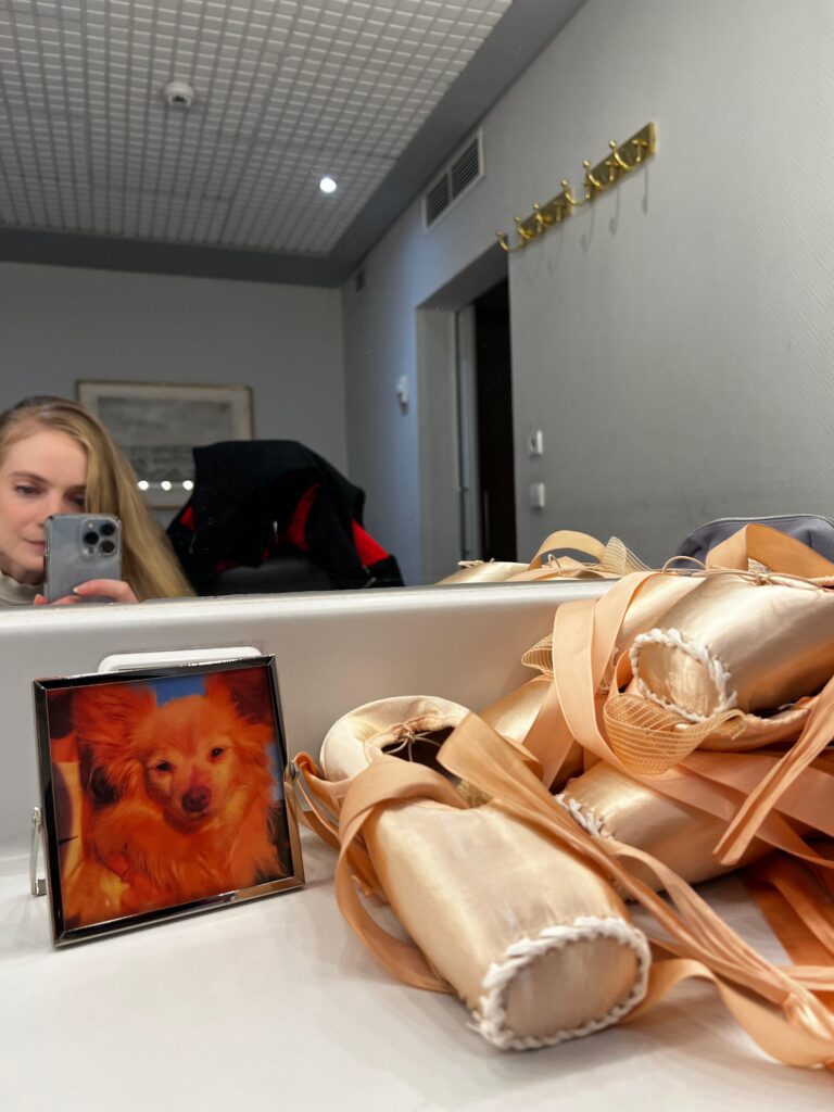 A young woman takes a selfie at her dressing room mirror, showing several pairs of darned pointe shoes and a small framed photo of her dog on the counter.