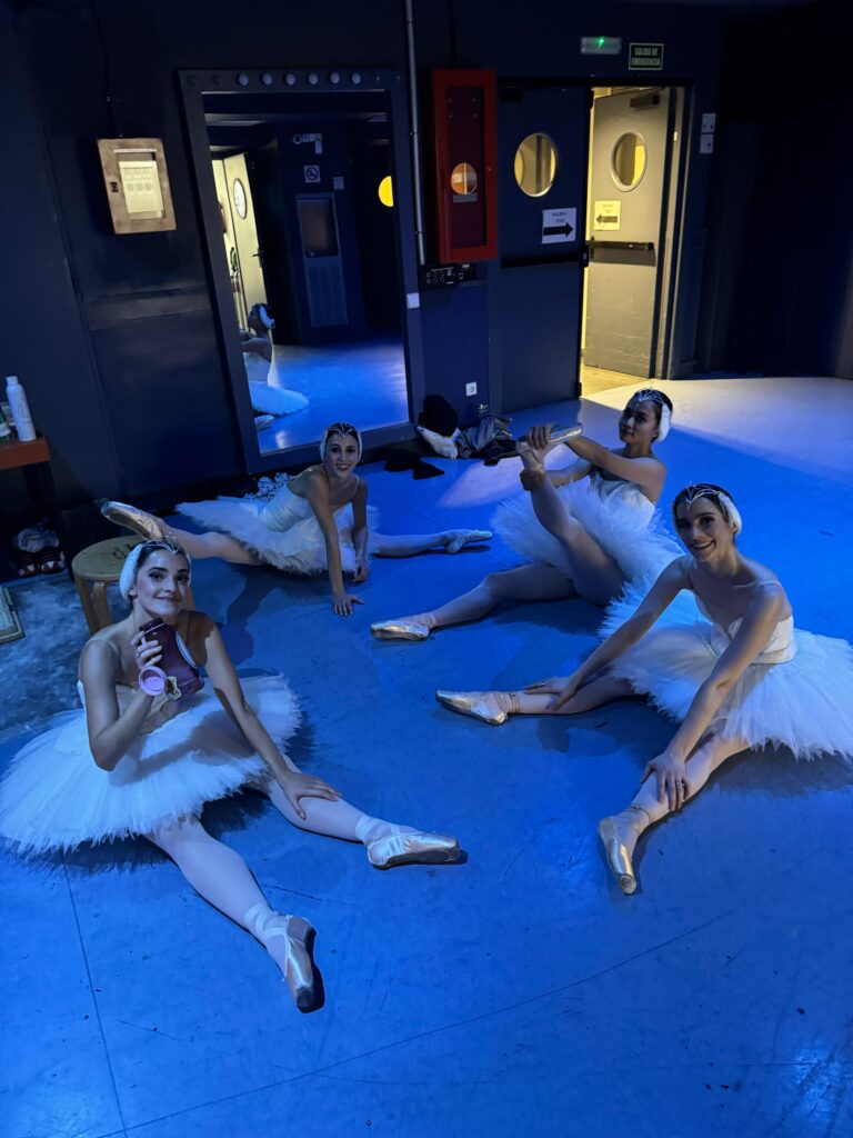 Four female dancers in white tutus, pink tights, pointe shoes and white feathered headpieces sit on the floor backstage and stretch.