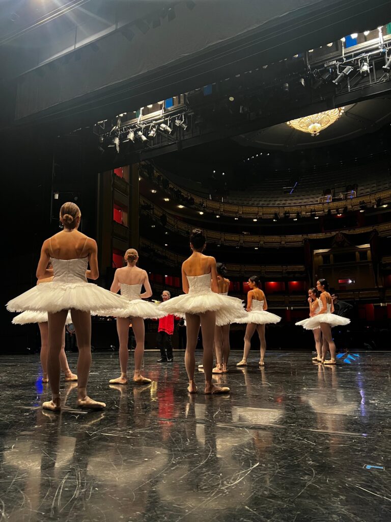 Eight young ballerinas in white tutus, tights and pointe shoes are shown from behind, standing onstage as a female rehearsal director gives notes.