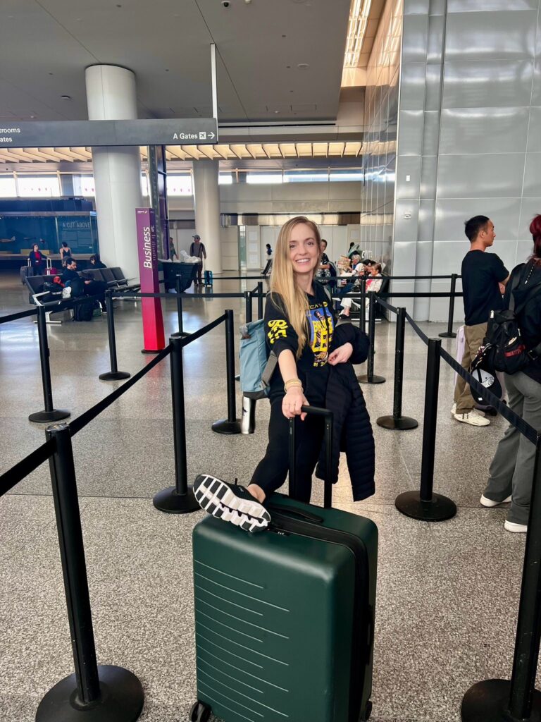 A young woman with long blonde hair props her right foot on top of her reen suitcase while waiting in line at airport security. She wears a black long-sleeved t-shirt and leggings and smiles towards the camera.