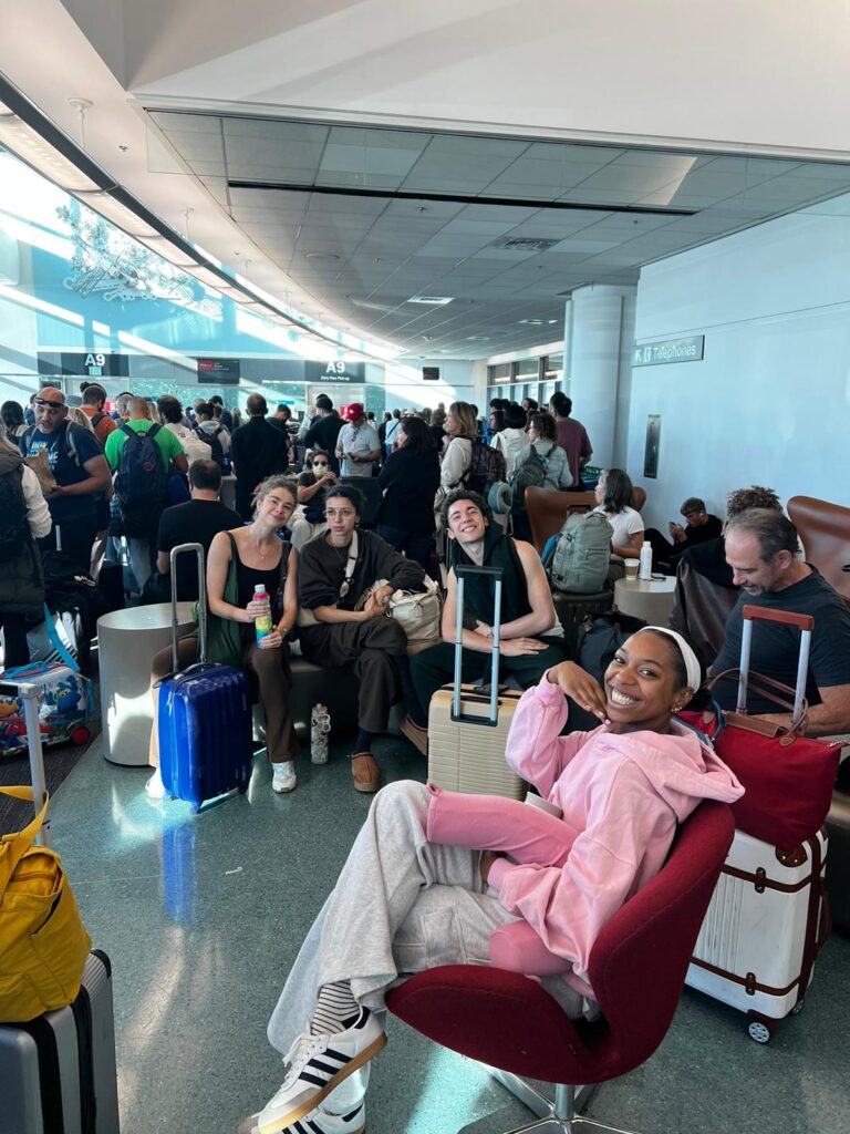 Four friends in casual clothing sit at a crowded airport, their suitcases nearby, and smile towards the camera.