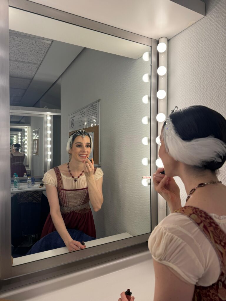 A female ballet dancer applies lipstick at her dressing room mirror. She wears a white feathered headpiece and a brown peasant dress costume.